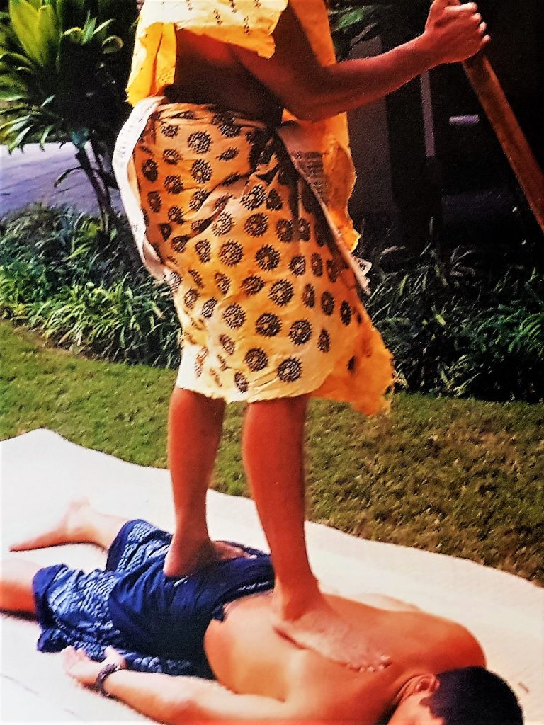 A lomilomi wāwae practitioner is walking barefoot on the back of his clilent. One footis on the sacrum, the other foot walks upwards along the spine. The client is lying down on the ground on a hala mat.