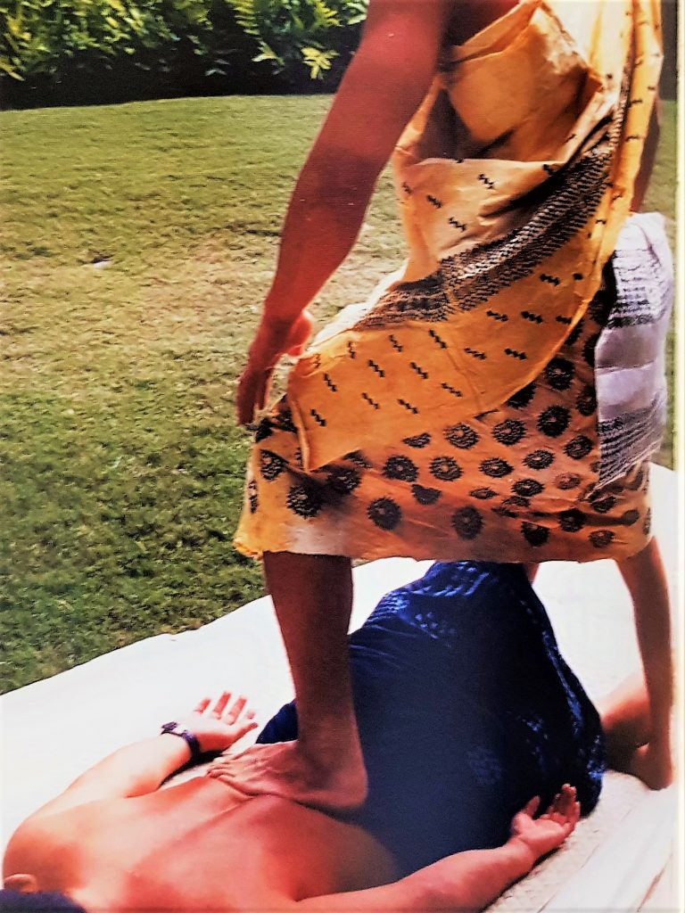 A lomilomi wāwae practitioner working with his feet onthe lower back of his client. The client is lying down on a hala mat on the floor.